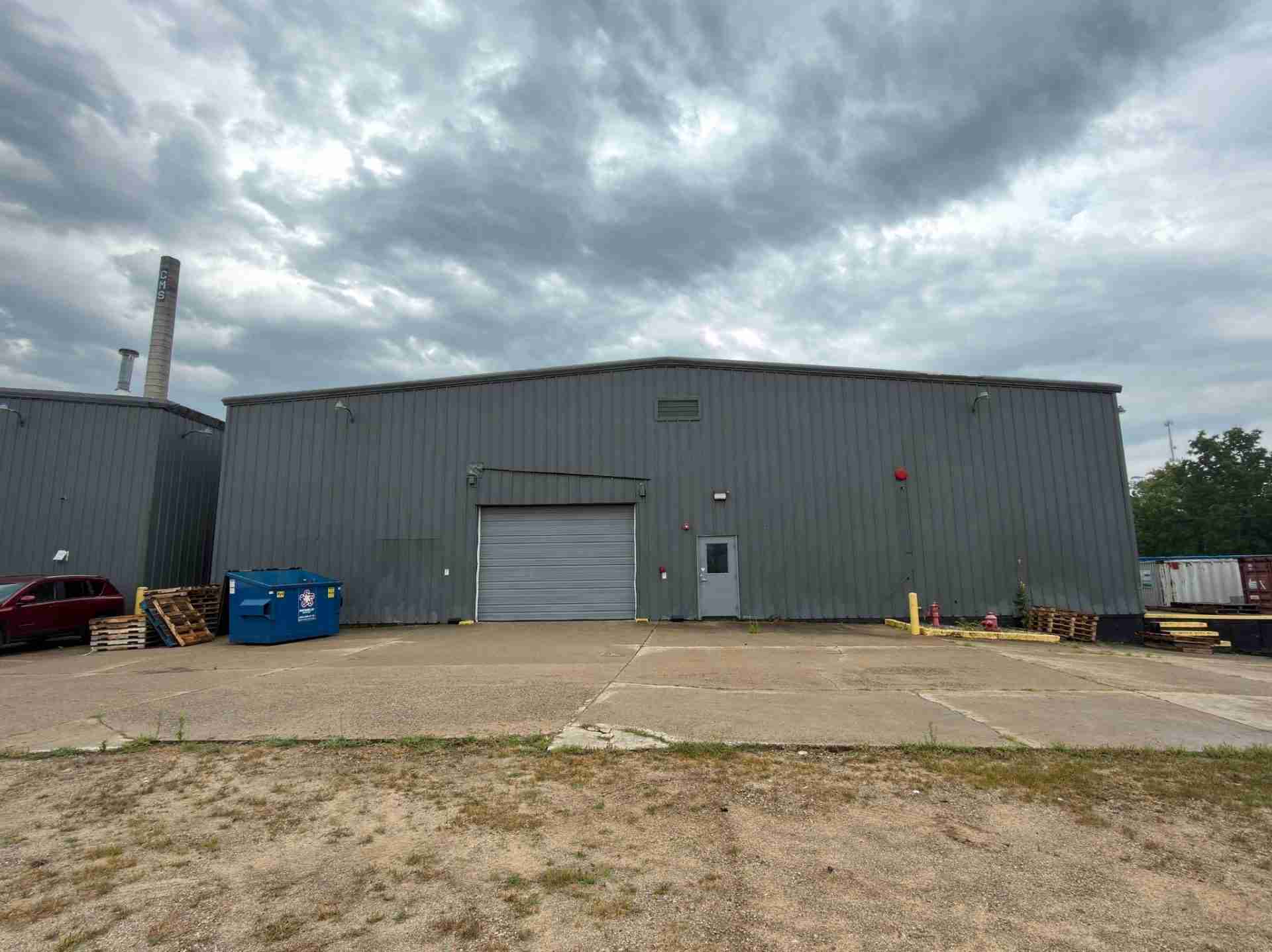 Industrial warehouse exterior with gray metal siding, a tall chimney, loading bay doors, and pallets/dumpster near the building under a cloudy sky.