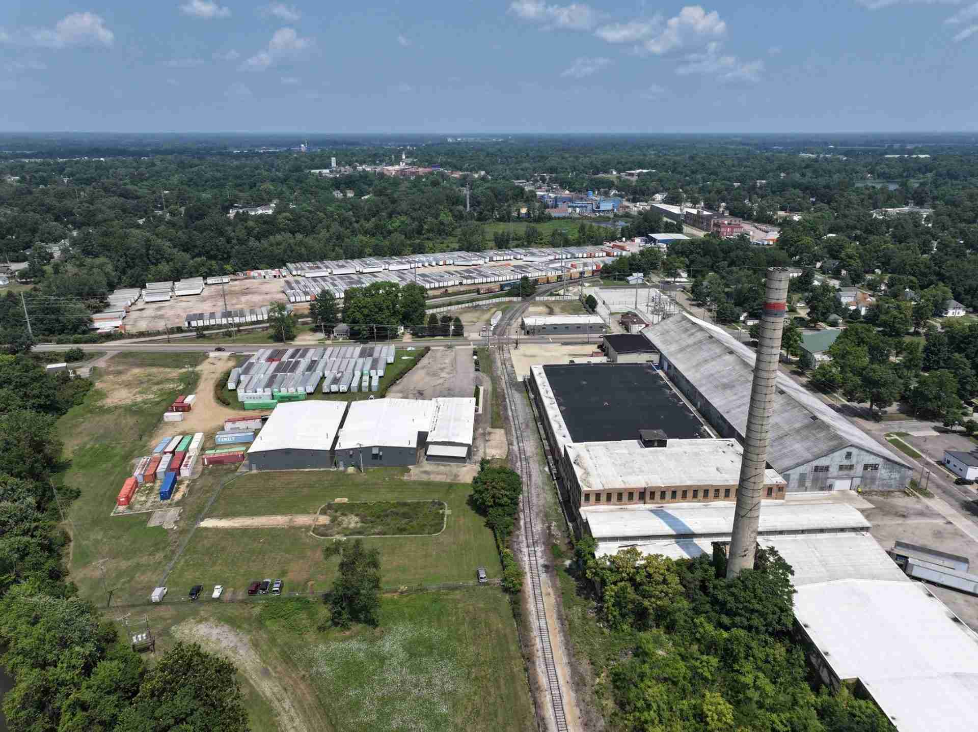 Aerial view of an industrial yard with rows of shipping containers, rail tracks, and large factory buildings near a forested area.
