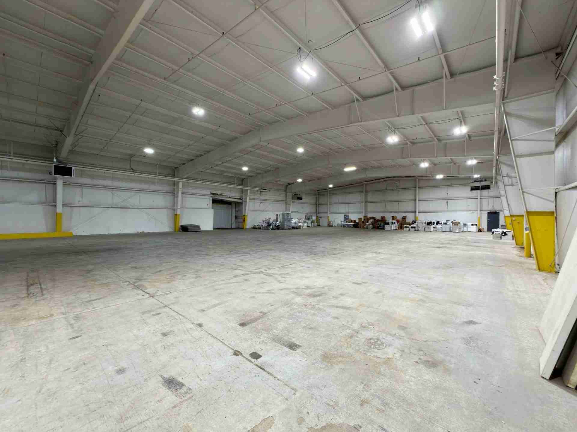 Spacious empty industrial warehouse with high white ceiling, bright overhead lights, and pallets stacked along the far wall. Yellow safety barriers flank columns by the walls.