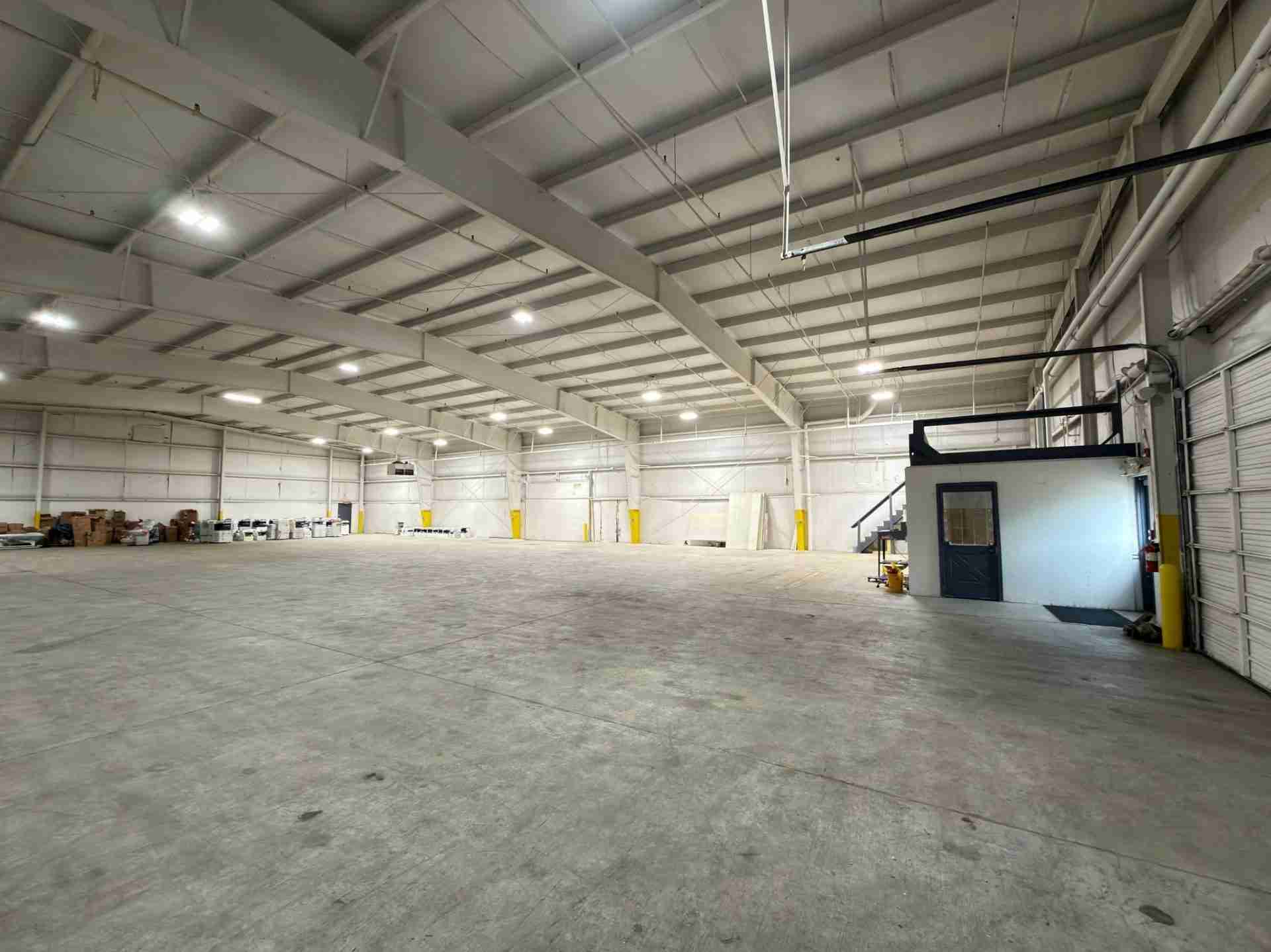 Interior of a large warehouse with exposed metal beams, bright lights, and a small office booth on the right; pallets stacked along the back wall.