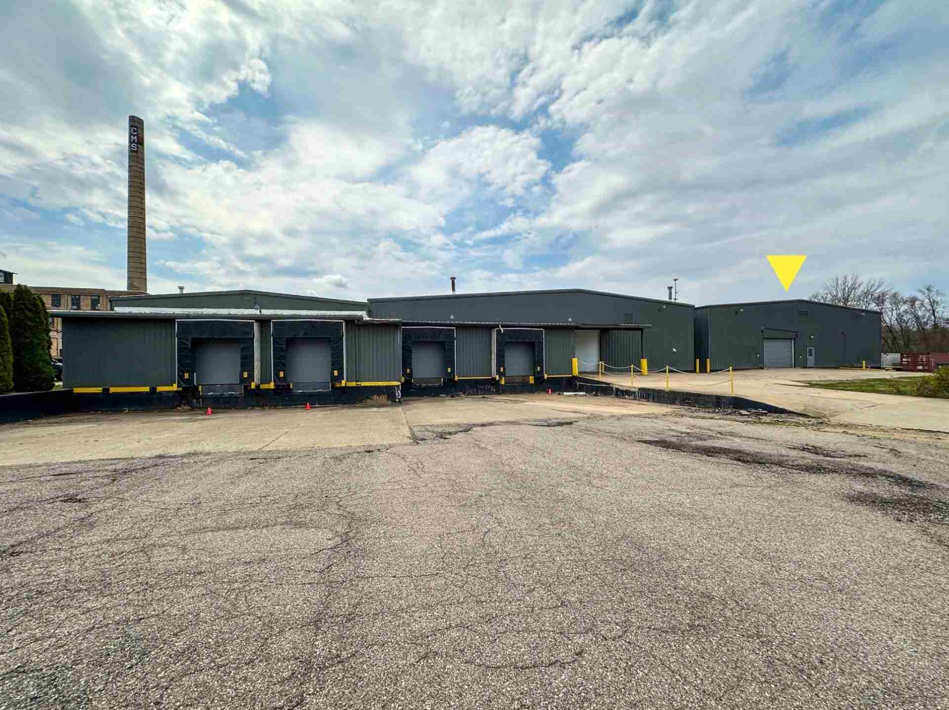 Industrial loading dock building with multiple bays, a tall CMS smokestack, and a cloudy sky.